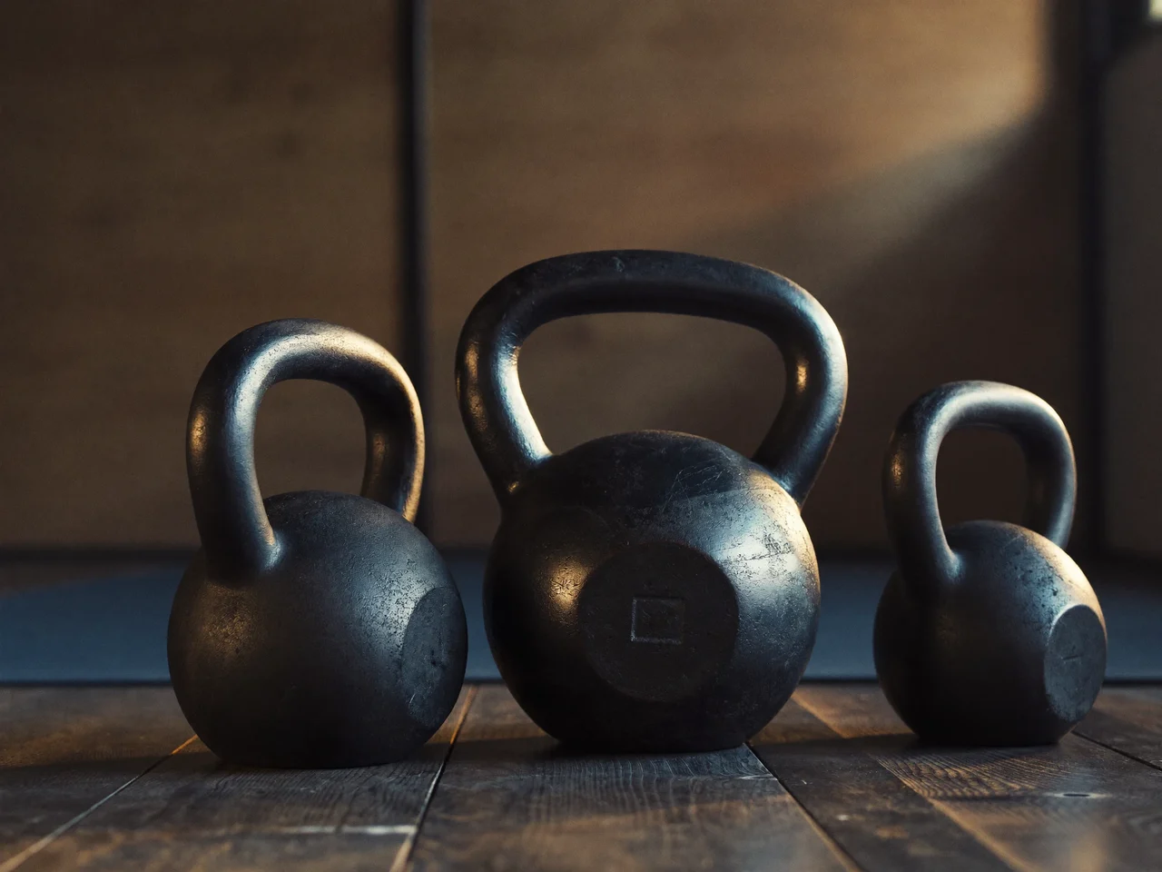 Tre kettlebell in ghisa di taglie diverse su un pavimento di legno consumato.